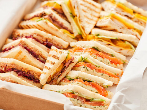 A variety of different triangle-cut sandwiches are presented in a box lined with white paper. Fillings include salmon, beef, cheese, and cranberry. Perfect catering food