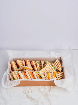 A variety of grilled sandwiches, cut into triangular portions, are packed inside a cardboard catering box lined with white paper, sitting on a marble-like table