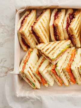 Rectangular box is filled with triangles of sandwiches that have grill marks. Sandwiches include peanut butter and jelly, salmon and cucumber, and various deli meats