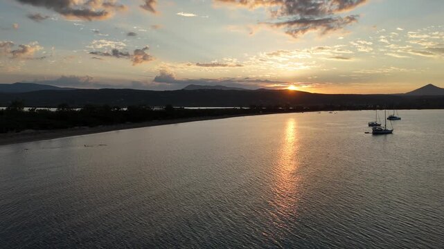 Navarin,Messinia,Aerial birds eye view forward from Giolova Beach towards Gialova Lagoon during sunrise.Beautiful panoramic scenery of mountains,reflection of the sun over sea where sailboats anchored