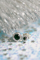 Handmade mini silver heart-shaped stud earrings with green gemstones on a Christmas sparkly background.