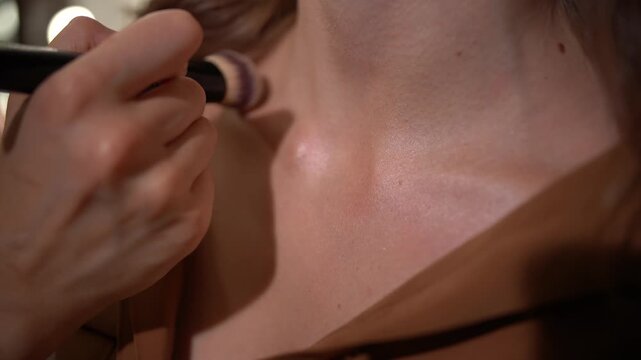 Makeup artist applying highlighter on woman's neck and decolletage with brush.