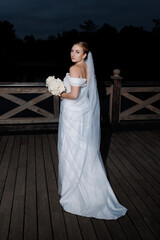 Beautiful bride poses at dusk on a wooden deck holding a bouquet, ready for a romantic outdoor...