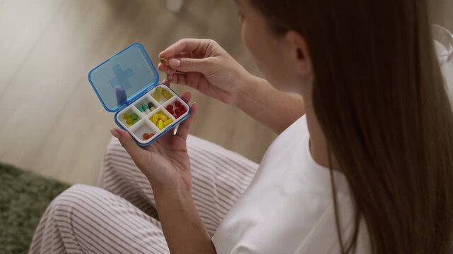 Young Caucasian woman in cozy homewear sitting on sofa as opening blue plastic pillbox with colorful capsules and taking her daily supplements, drinking them with glass of water for healthy routine