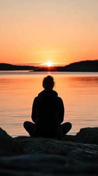 Meditative figure sits peacefully on rocks, observing a glorious sunset over serene water, evoking spiritual devotion and tranquil contemplation.