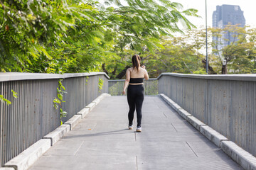 A confident plus-size Asian woman jogs along an outdoor urban walkway surrounded by greenery, enjoying an active lifestyle.