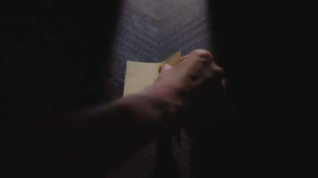 A close up of "Saturday" being written on a Post it Note. The note is then picked up and placed back down. Shot in dramatic lighting and slow motion.