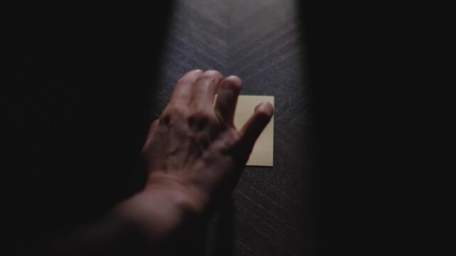 A close up of a hand writing "Tuesday" on a Post it Note with a marker. The hand picks up the paper and places it back down in the light. Shot in Slow motion.