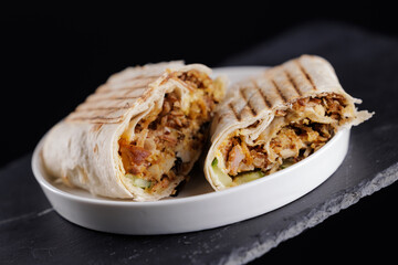 Grilled chicken shawarma wrap with vegetables on white plate on dark background