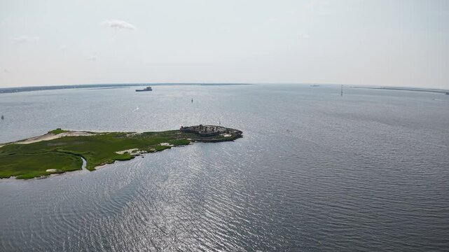 Drone aerial footage capturing a solitary patch of green land surrounded by expansive blue water and distant boat near Charleston harbor, South Carolina.