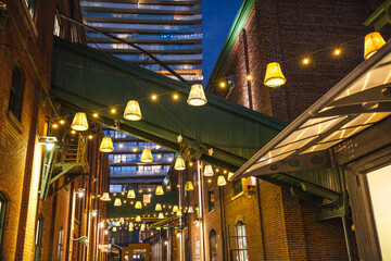Naklejka premium Decorative hanging lamps and warm string lights illuminating a narrow brick alley in Torontos Distillery District at night.