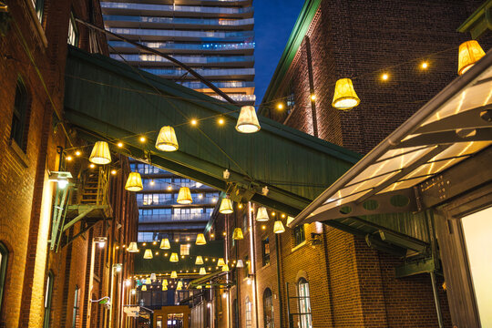 Decorative hanging lamps and warm string lights illuminating a narrow brick alley in Torontos Distillery District at night.