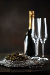 Black caviar and champagne. An exquisite snack. Glasses and a bottle of champagne are out of focus. Black caviar is served in a crystal dish with ice. The concept of a festive treat.