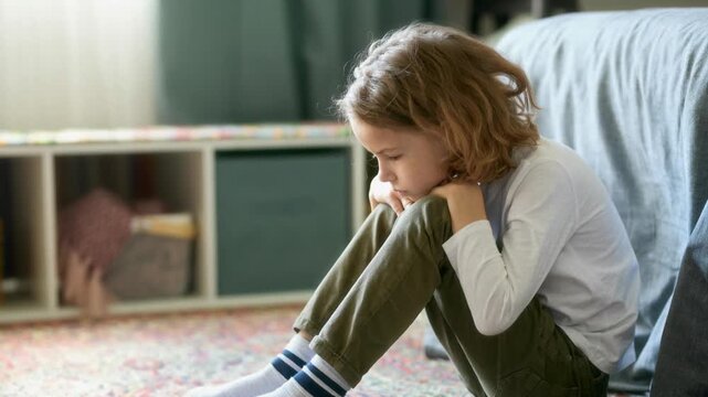 Side view of upset boy child sitting on floor holding knees while regulating feelings in bedroom, AI generated footage
