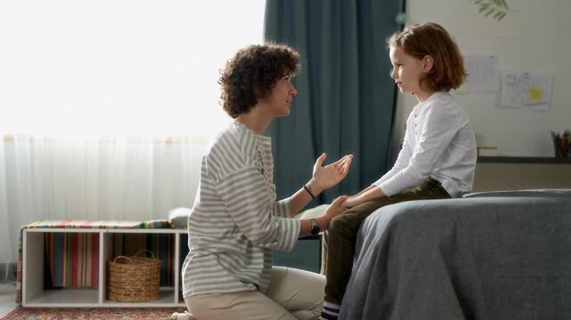 Side view of mother comforting upset young son gesturing while discussing feelings together in bedroom, AI generated footage