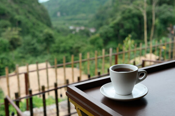 A warm cup of coffee placed on a wooden table with a serene green mountain view, creating a calm and relaxing atmosphere