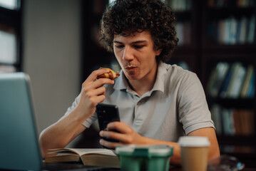 Young man eating and using smartphone in library or home office