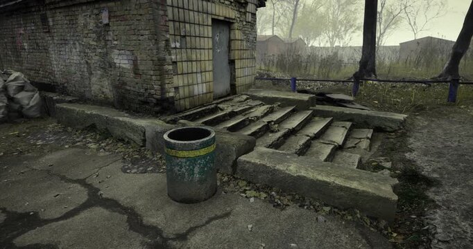 An abandoned location displays a worn trash bin on cracked pavement, with overgrown grass and trees surrounding crumbling structures. Nature gradually takes over this forsaken area.
