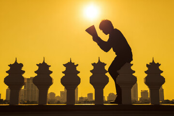 Silhouette man reading book at sunset on rooftop with city skyline and decorative balustrade, contemplative warm light