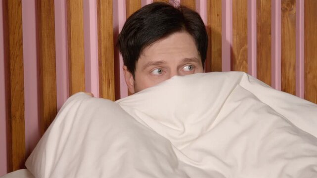 Scared attractive bearded middle aged man suffering from insomnia, covering half face and peeking out from blanket, looking at sides with frightened expression while lying in bed at night.