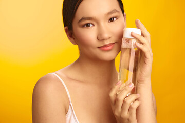 asian woman with dark hair wearing a light top holds a transparent bottle against a bright yellow...