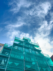 Bright turquoise scaffolding envelops a building under a vast blue sky with fluffy clouds in a vibrant city landscape