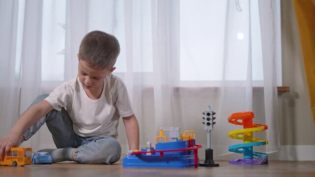 Preschooler boy plays with toy bus. Child sits on floor near toy garage. Preschooler moves bus past toy cars. Boy enjoys vehicles. Toy bus drives around learner playset. Child, boy, preschooler.