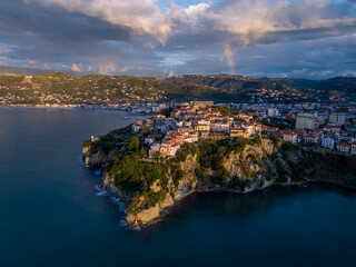 Aerial view of a promontory crowned with ancient architecture, kissed by the golden light of the setting sun, rainbow arcing above, Agropoli, Campania, Italy.