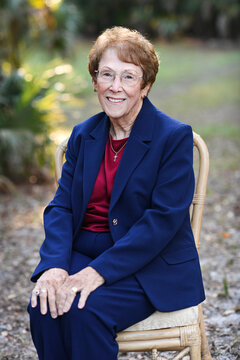 Smiling elderly woman seated outdoors in a navy blue suit.
