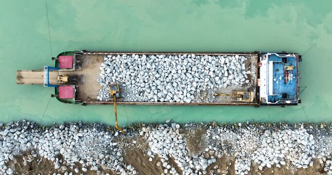 Top-down aerial view of an excavator on a barge unloading large riprap stones to build a coastal breakwater in turquoise water, a cinematic 4k footage of industrial engineering and construction.