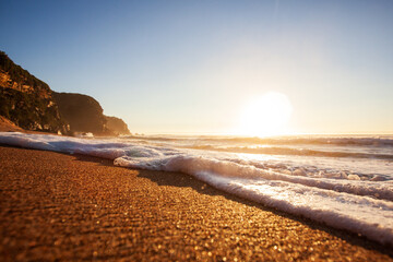 Close-up view of a foamy wave rolling up a golden textured sandy beach as a bright sun rises above the ocean. Captured from a low perspective on Stanwell Park Beach on the Illawarra South Coast.