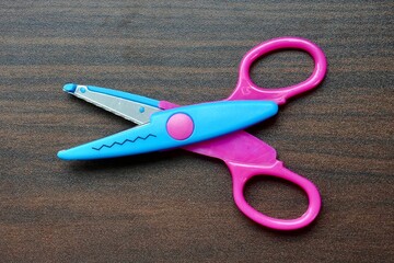Pink and blue colored zig zag craft tool scissors object isolated on horizontal brown table surface background from top above angle view.