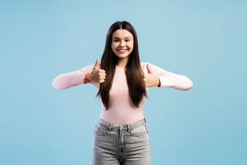 Happy smiling teen girl giving double thumbs up gesture looking at camera posing on blue background