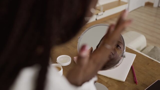 Over-the-shoulder shot of young Black woman applying hydrogel eye patches while looking in mirror on wooden countertop during her morning skincare routine in bright modern apartment