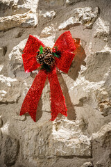 Red glitter Christmas bow with pine cones and evergreen accents mounted on stone wall, festive winter decoration outdoors, holiday ornament detail, seasonal decor concept, rustic Christmas atmosphere
