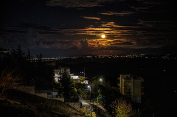 Night cityscape with full moon rising above clouds, dramatic sky, glowing urban lights, quiet hillside neighborhood, moody atmosphere, long exposure landscape, serene nocturnal scenery