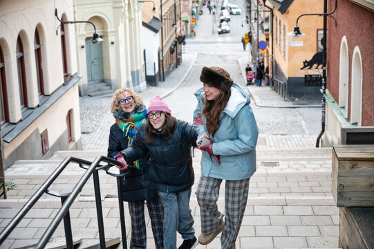 Three Caucasian women smiling and climbing stairs in Stockholm on a winter day, one with cerebral palsy supported by friends, enjoying urban life and friendship in a cheerful outdoor atmosphere