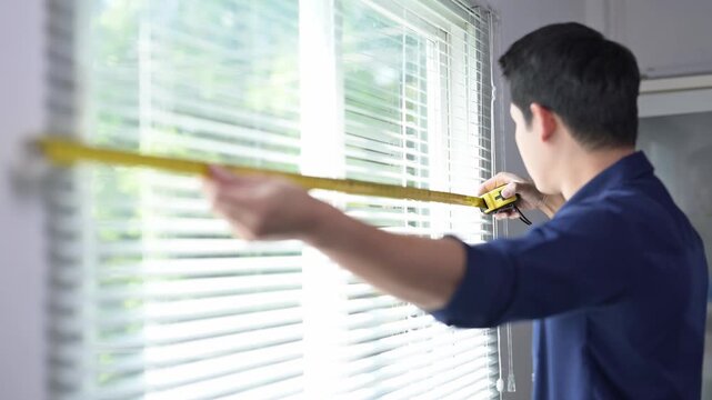 A man is measuring a window with a tape measure. He is wearing a blue shirt. The window is white