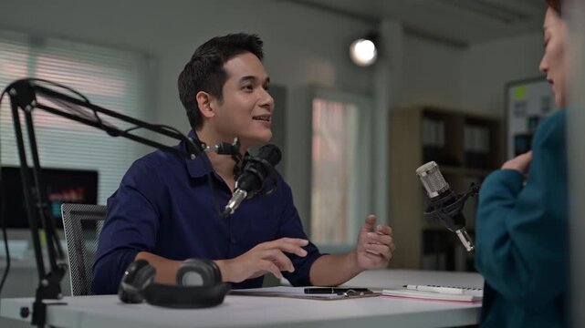 A man is talking into a microphone. A woman is sitting behind him. There are two microphones on the table