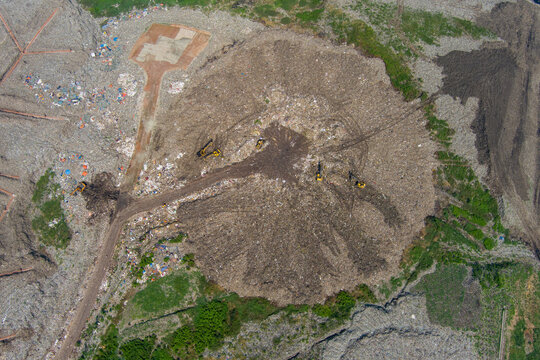 Aerial view of a vast landfill where heavy machinery sculpts the landscape of waste, creating an stark contrast against the surrounding greenery, Dhaka, Dhaka Division, Bangladesh.
