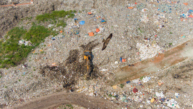 Aerial view of a vast landfill where colorful refuse contrasts with the dark earth turned by machinery, while a bird soars above, Dhaka, Dhaka Division, Bangladesh.