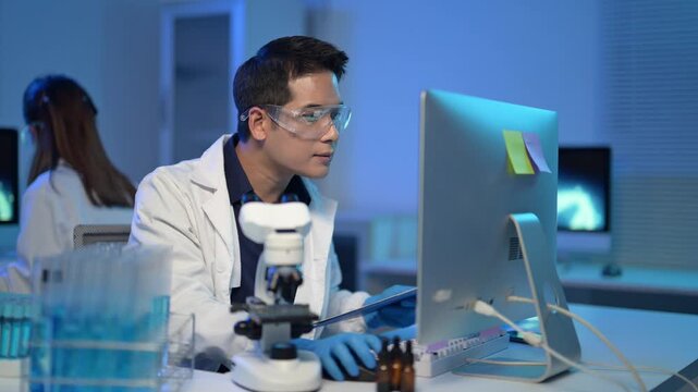A man in a lab coat is looking at a computer monitor. There are several bottles on the table, and a person is sitting behind the man