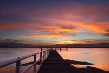 Sunset fills the sky with colour with beautifully vibrant colours over a jetty in a saltwater lake. Captured at Boonerah Point looking west to the mountains on Lake Illawarra, NSW, Australia.