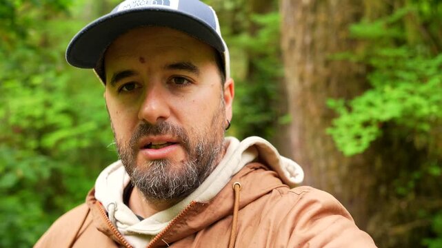 Caucasian bearded man wearing a baseball cap and a brown jacket films himself talking in a lush green forest. Likely creating content for social media or a vlog about his travels on vancouver island