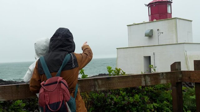 Tourists wearing raincoats and backpacks pointing at the ocean near amphitrite point lighthouse in ucluelet, vancouver island, british columbia, canada, on a cloudy day