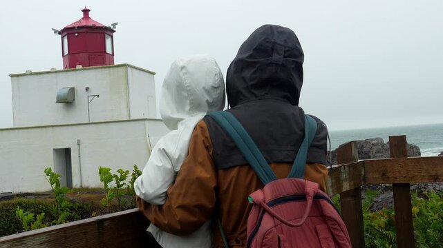 Parent and child, wearing raincoats and hoods, stand at a wooden railing, gazing at the amphitrite point lighthouse on a misty day in ucluelet, vancouver island