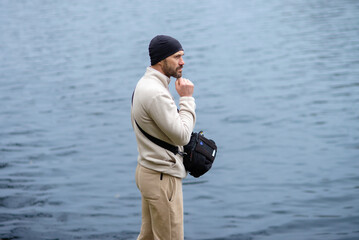 Thoughtful man in a beanie and fleece jacket standing by calm water, contemplating or observing. Ideal for themes of solitude, nature, travel, or reflection. © dizfoto1973