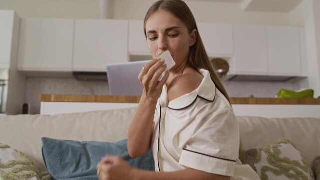 Young Caucasian woman sitting on comfortable couch and using transdermal patches on different body parts as part of her wellness routine at modern minimalist-style home setting