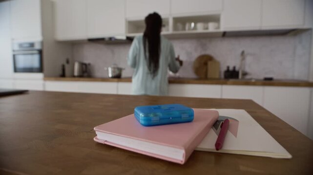 Young Black woman reaching for blue plastic pillbox on notebook at wooden table in bright minimalist kitchen with calm morning atmosphere