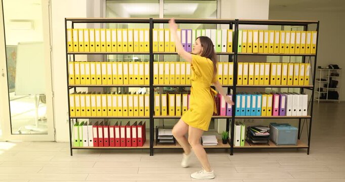 Joyful woman office worker in bright yellow dress dances energetically in modern office. Cheerful female moves to imaginary rhythm against shelves